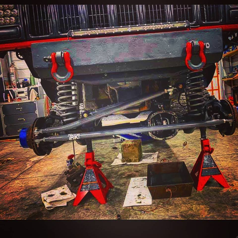 BKFabworks high-clearance grey steering kit installed on lifted Jeep front axle with coil springs and red jack stands in shop setting