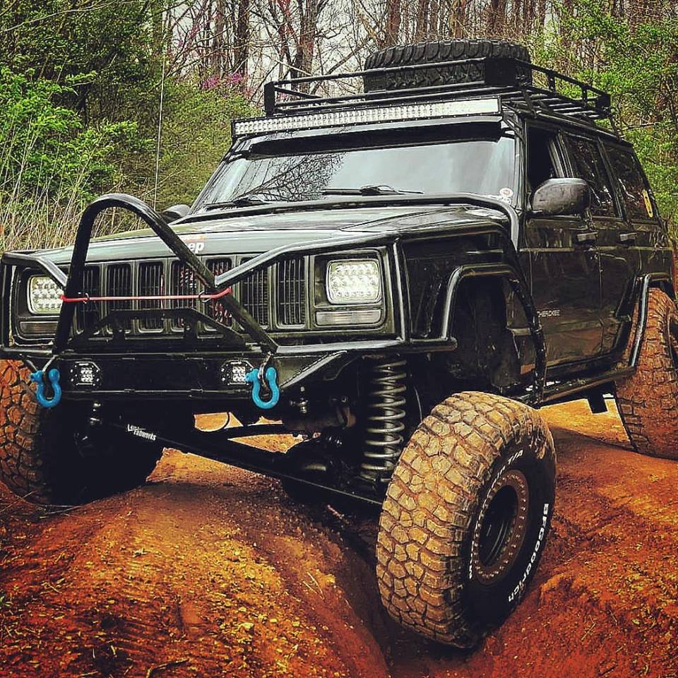 Black Jeep Cherokee XJ with upgraded suspension flexing in a trail rut, showing off-road clearance and BKFabworks components including steering and bumpers