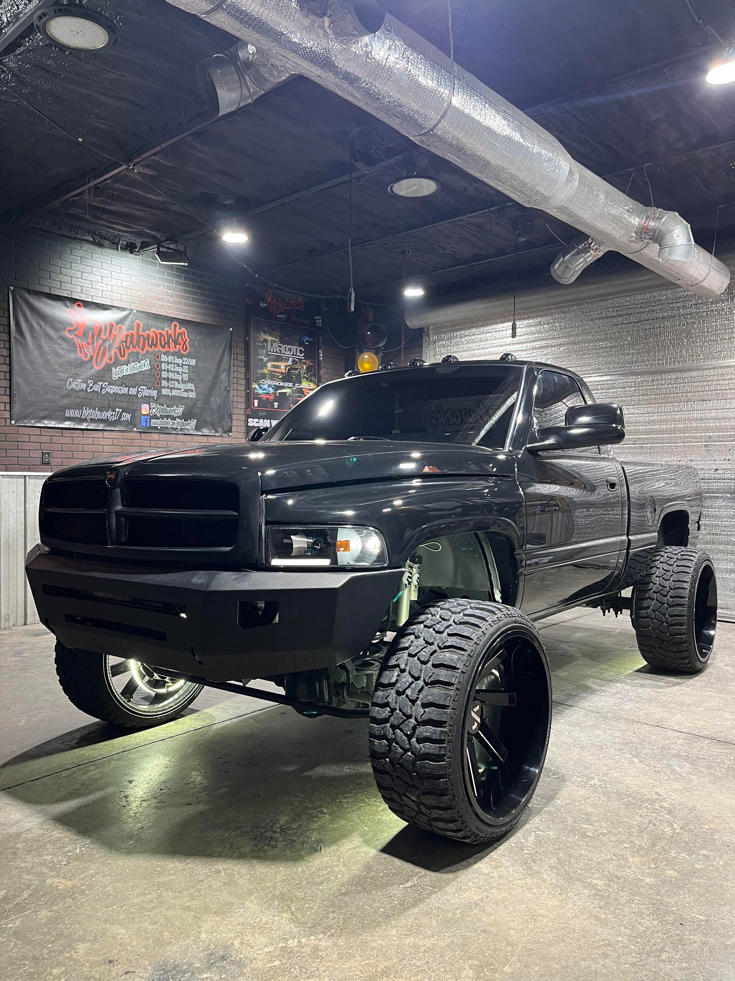 Black lifted 94–02 Dodge Ram with aftermarket wheels and suspension parked inside BKFabworks shop with custom lighting and branding